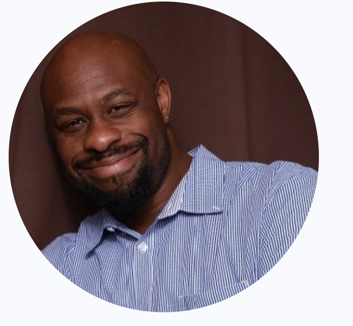 
A bald, African- American man with a beard is smiling at the camera. He is wearing a blue and white striped button-down shirt. 